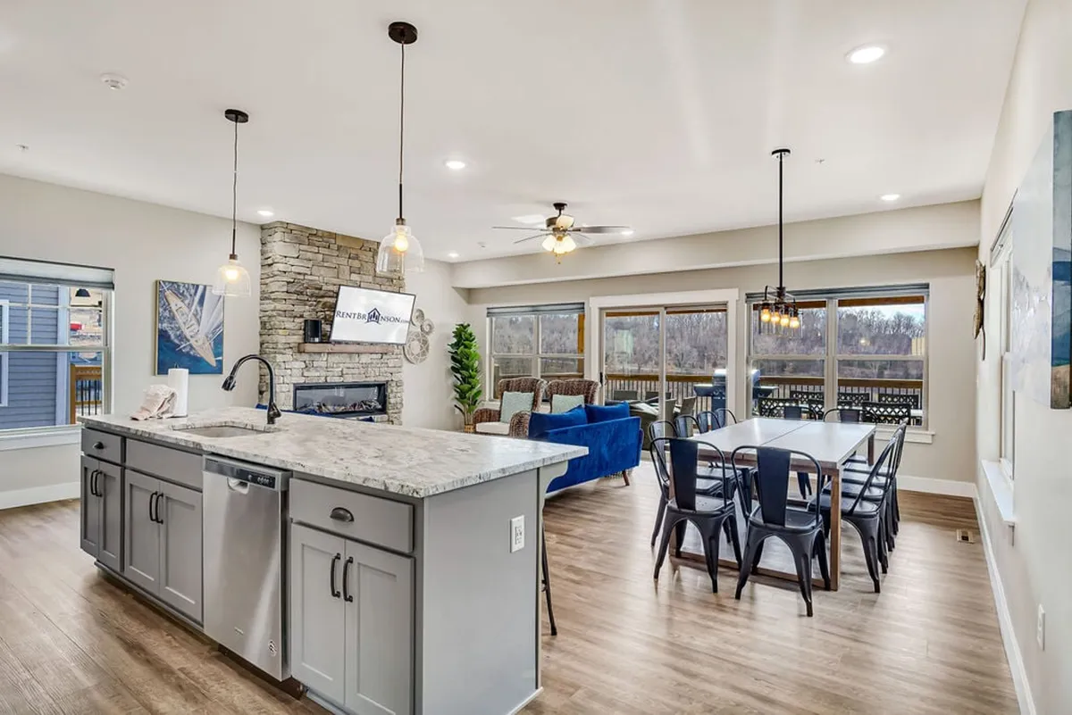 This image presents a harmonious open-concept space featuring a granite kitchen island, a stone fireplace, a large dining table, comfortable seating, and expansive windows. The scene captures the kitchen, dining, and living areas flowing seamlessly together, bathed in natural light.