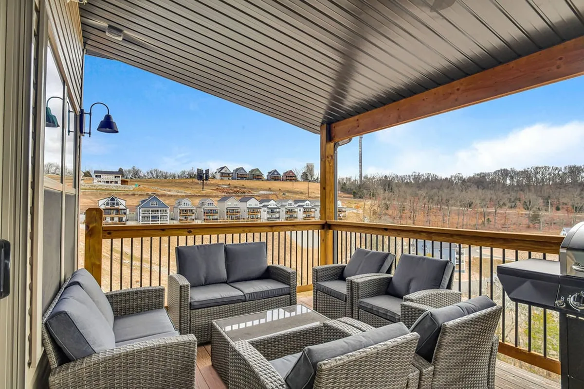 An outdoor seating area on a spacious deck overlooks rolling hills and neighboring homes. A grill is visible at the side, with comfortable chairs arranged for relaxation or dining, all set against a warm sunset glow.