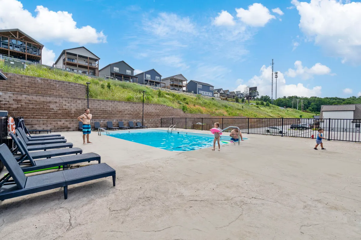 This image captures a lively outdoor pool scene with children playing in the water, families lounging on poolside chairs, and background houses adding to the neighborhood feel. The joyful atmosphere reflects summer fun and community togetherness.