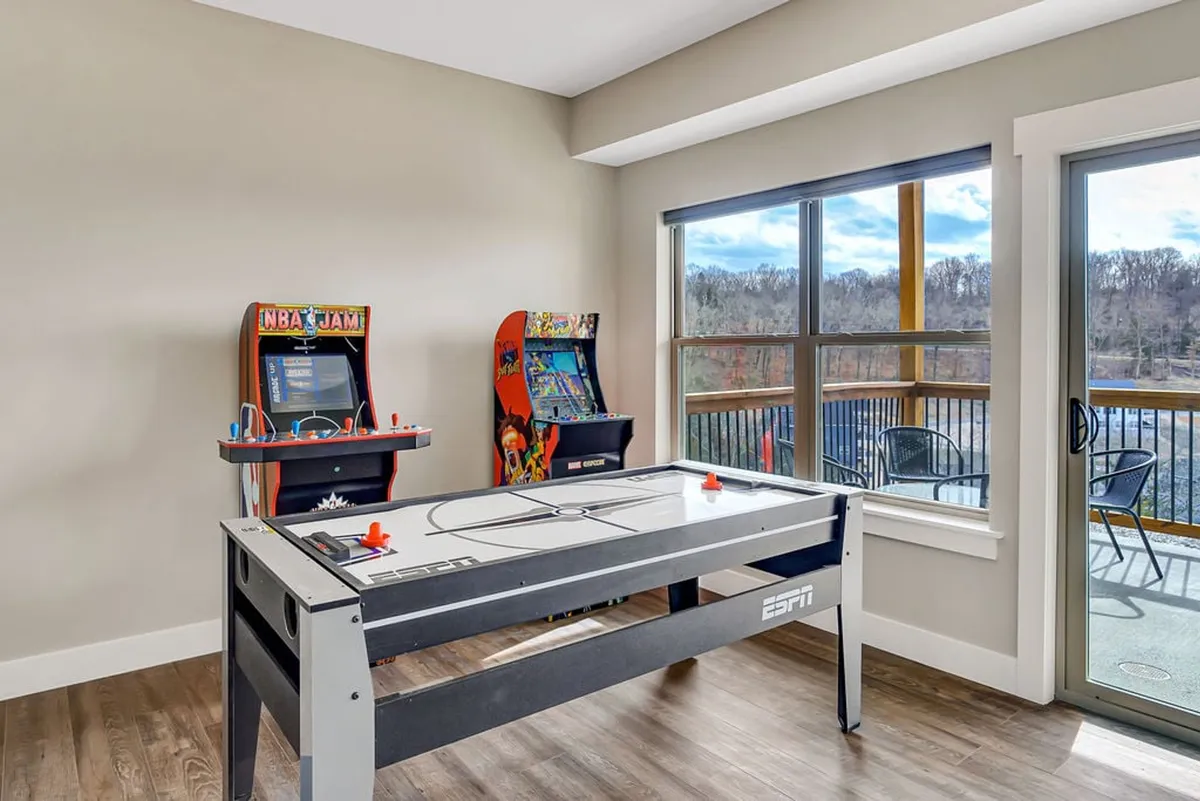An inviting interior game room equipped with an air hockey table, arcade machines, and large windows that let in natural light. Friends are pictured playing and socializing, with a scenic view visible through the windows.