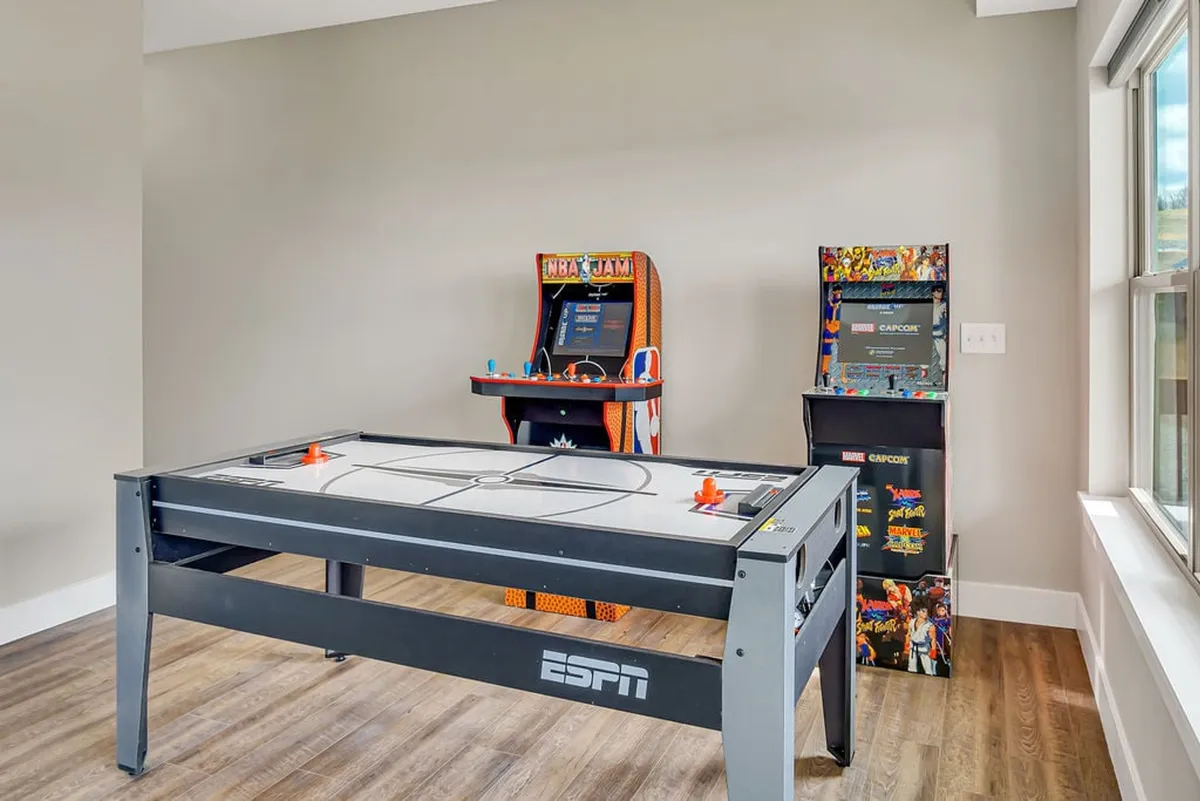 This energetic image captures the heart of the game room with an air hockey table as the centerpiece, flanked by classic arcade game cabinets (including NBA Jam and Capcom). The image shows a lively, colorful atmosphere perfect for friendly competitions and group fun.