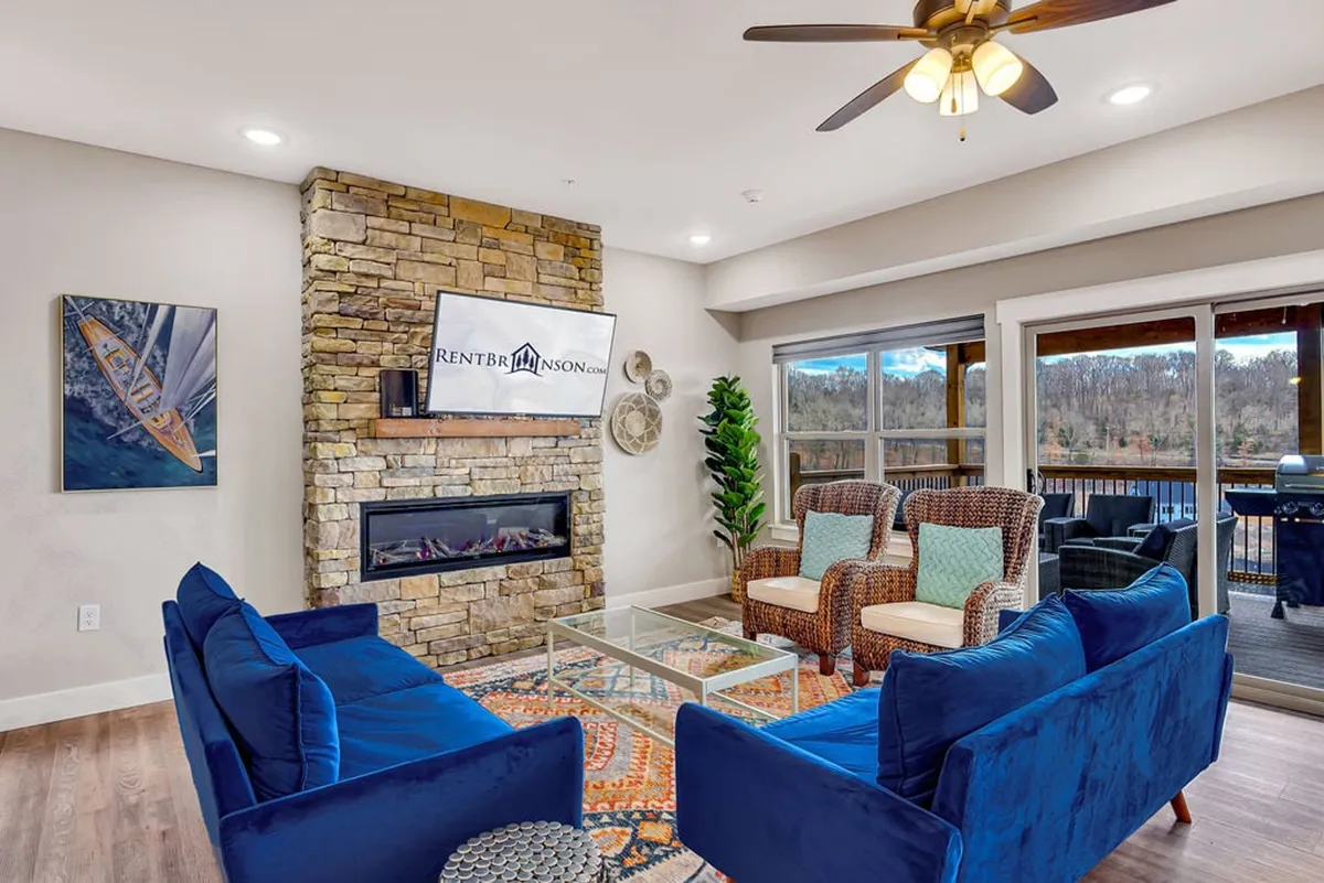 The image features a stylish, open-concept living room with plush blue sofas centered around a striking stone fireplace. Large windows allow natural light to pour in, highlighting the modern furnishings and artwork, creating a cozy and inviting environment for relaxation or gathering with friends and family.