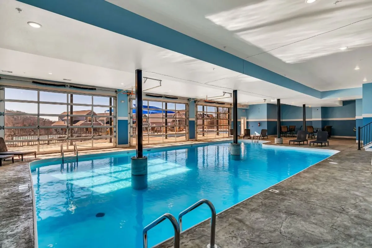 This shows an elegant indoor swimming pool with large windows letting in natural light and offering glimpses of the outdoors. Comfortable poolside seating provides space for relaxation and socializing in a resort-like, year-round setting.