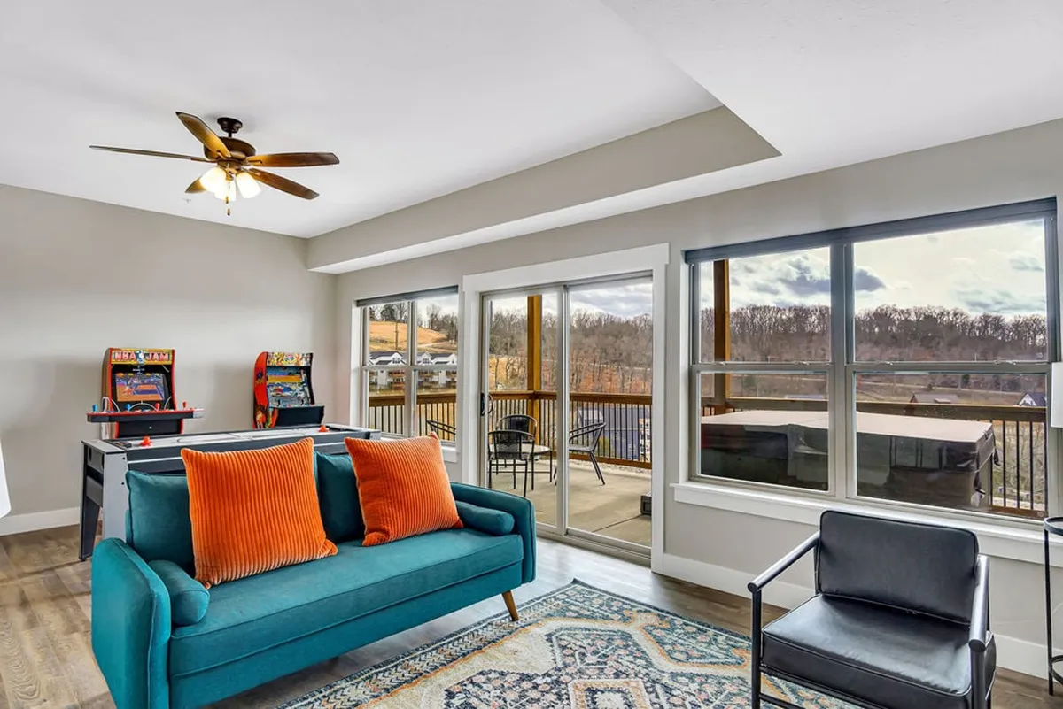 A spacious game room scene featuring a teal sofa with orange cushions, multiple arcade game machines, and large windows providing natural light and a view outside. The room feels relaxed and welcoming, perfect for both active play and downtime.