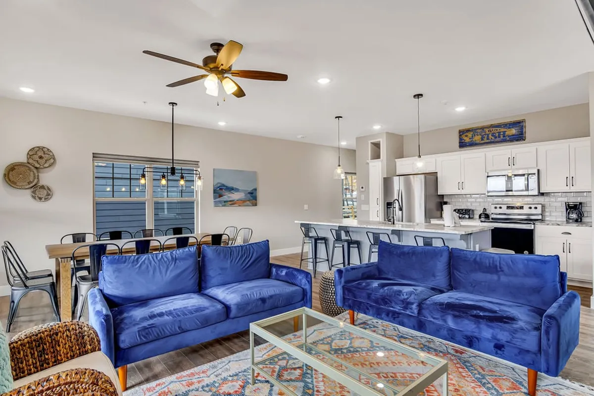 This scene captures the integration of the living area, dining table, and kitchen island in one spacious, inviting interior. Blue sofas, a glass coffee table, and a family-friendly dining table signal a space designed for both relaxation and lively meals.