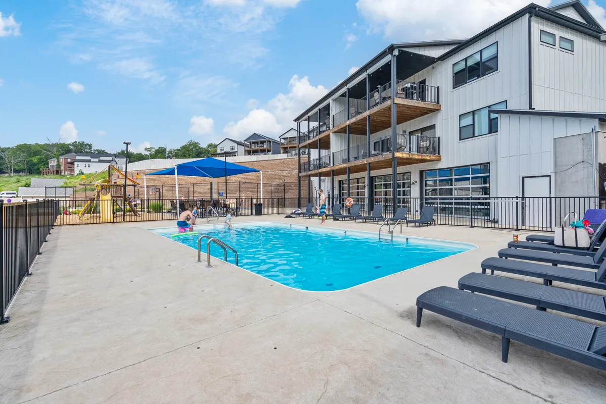 This exterior image captures a vibrant swimming pool area surrounded by lounge chairs and playground equipment, with balconies of the building visible in the background. People are socializing poolside while children play, evoking a sense of community and fun.