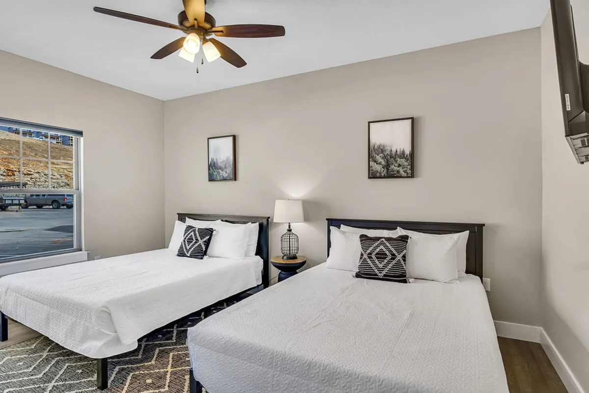 A cozy bedroom with two beds dressed in soft linens and decorative pillows, flanked by a bedside lamp and complemented by soothing wall art. A window allows daylight to brighten the room, creating a comfortable retreat for families or friends.