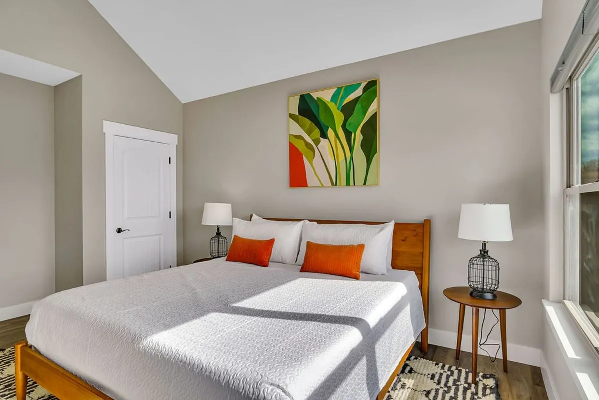 A spacious bedroom featuring a large bed with crisp white high-end linens, colorful artwork above the headboard, and nightstands with lamps. Natural light streams in from a nearby window, enhancing the tranquil and cozy atmosphere.