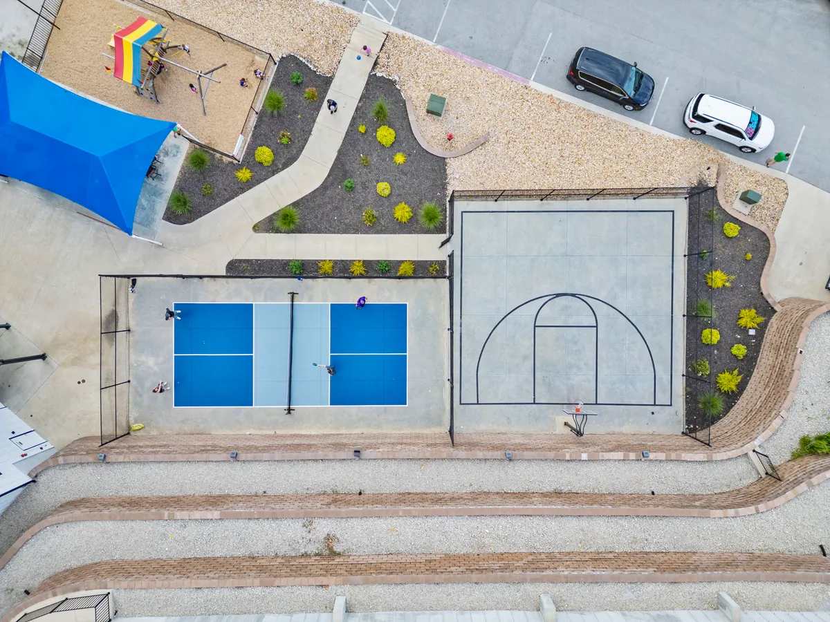 The image features expansive tennis and basketball courts, a playground area, and a blue shade structure set amidst landscaped gardens. Groups of people engage in games and socializing, while children play nearby.