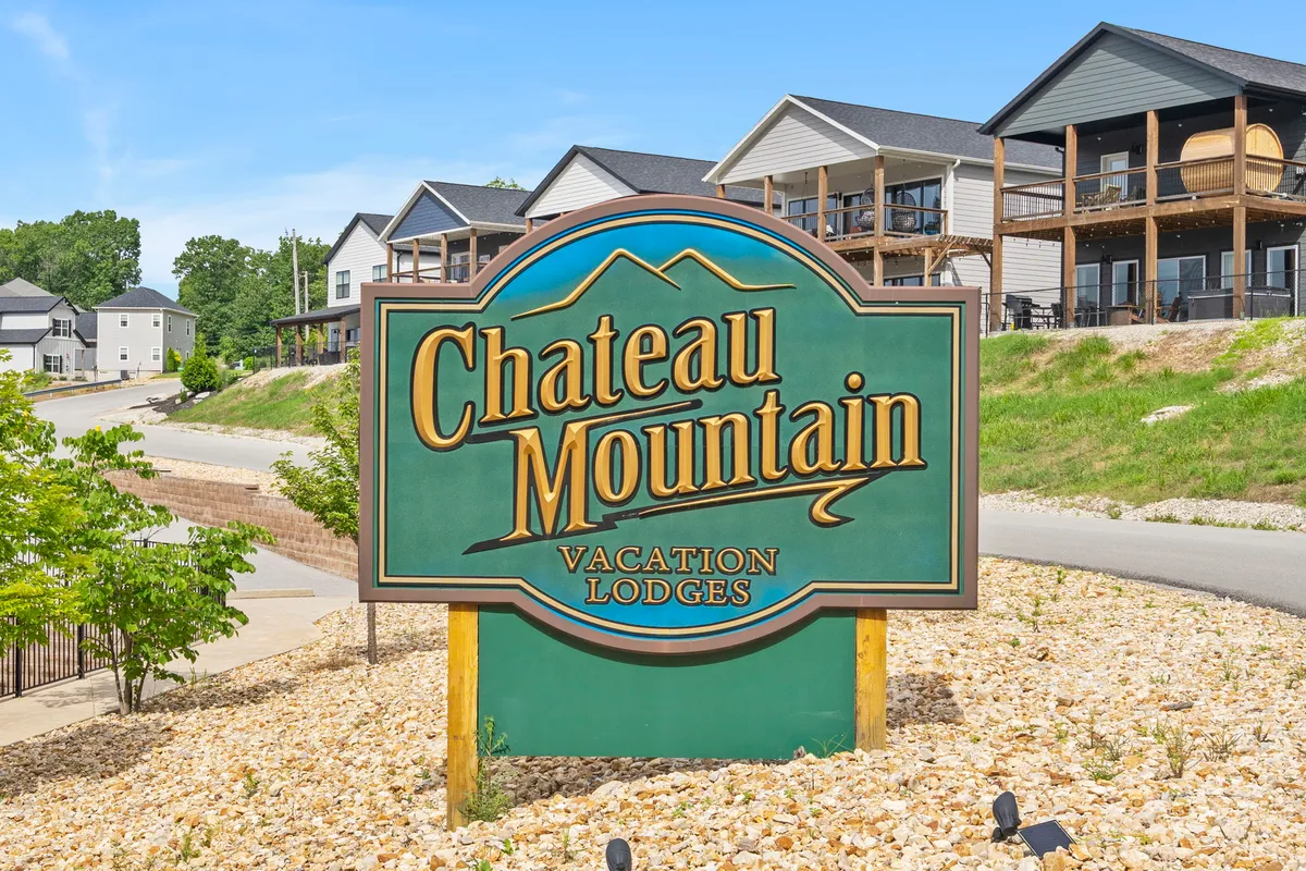 A wide exterior shot showcasing the Chateau Mountain sign at the entrance, surrounded by well-maintained landscaping and several inviting lodges set against a backdrop of rolling hills. The area exudes a sense of arrival and community, making it clear this is a destination for family gatherings and outdoor activities.